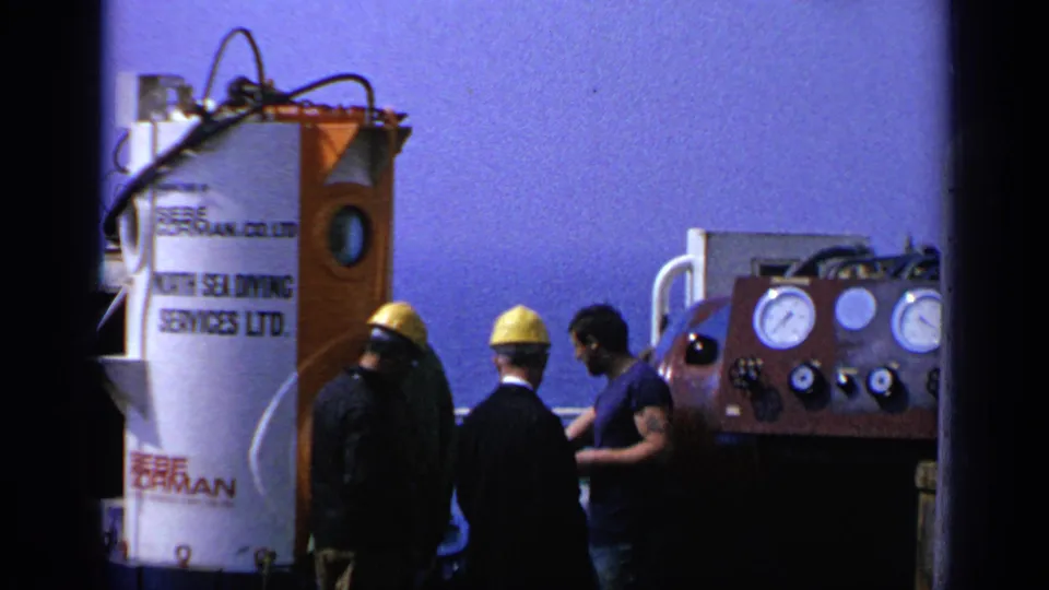 1970 United Kingdom shipboard frame showing men beside a diving chamber and a gauge panel on deck.