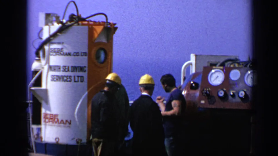 Workers on a 1970 United Kingdom vessel deck stand between a labeled diving chamber and pressure gauge equipment.