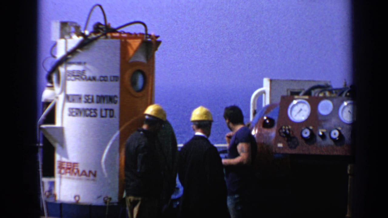 Workers in yellow hard hats stand beside a North Sea Diving Services Ltd. chamber and control panel.