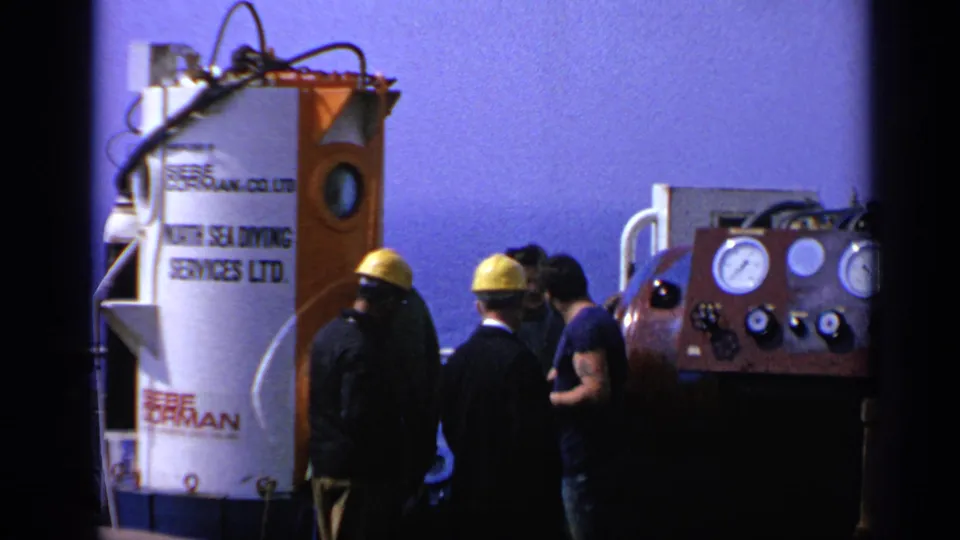 Workers at a 1970 UK offshore site appear to confer beside diving-support equipment and analog controls.