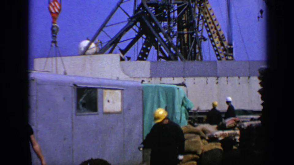 A 1970 UK offshore platform scene shows workers moving below a crane with deck structures, tarps, and materials visible.