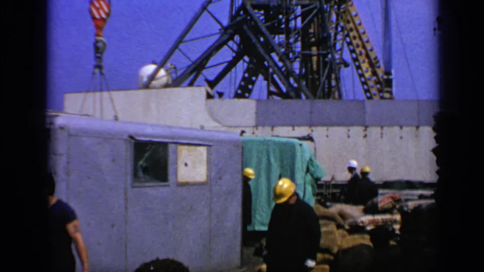 End frame from a 1970 UK offshore platform worksite with hard-hatted workers, crane elements, and construction materials.