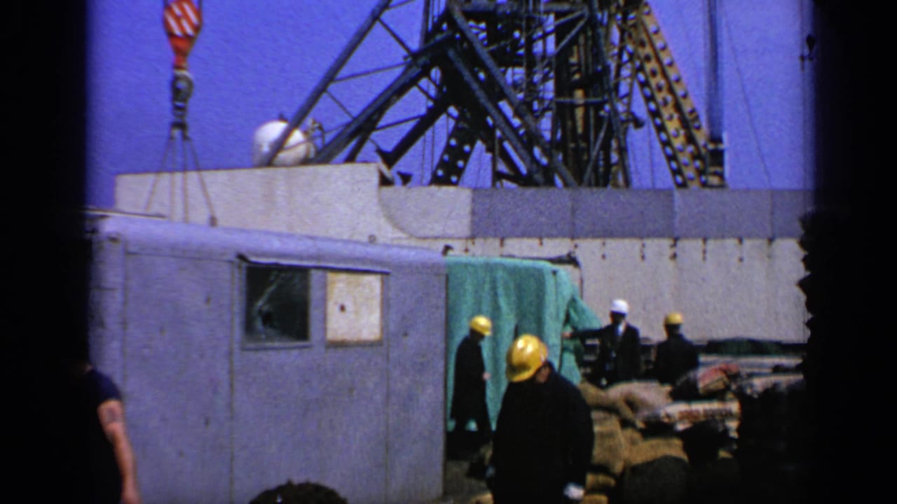1970 United Kingdom 8mm frame showing hard-hatted workers beside maritime platform equipment and a suspended crane hook.