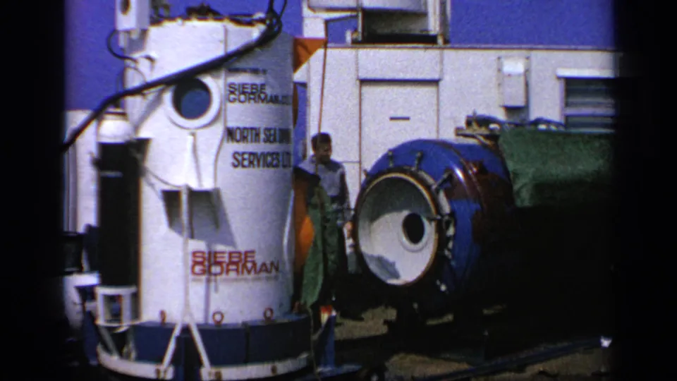 Closer 1970 United Kingdom view of a labeled Siebe Gorman cylinder with workers and a blue cylindrical module nearby.