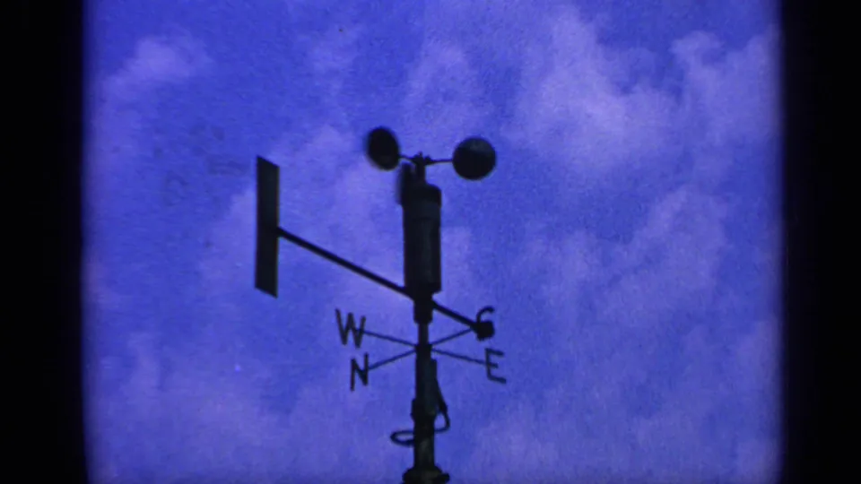 Weathercock with directional letters and spinning cups silhouetted against blue sky in the same 8mm maritime sequence.