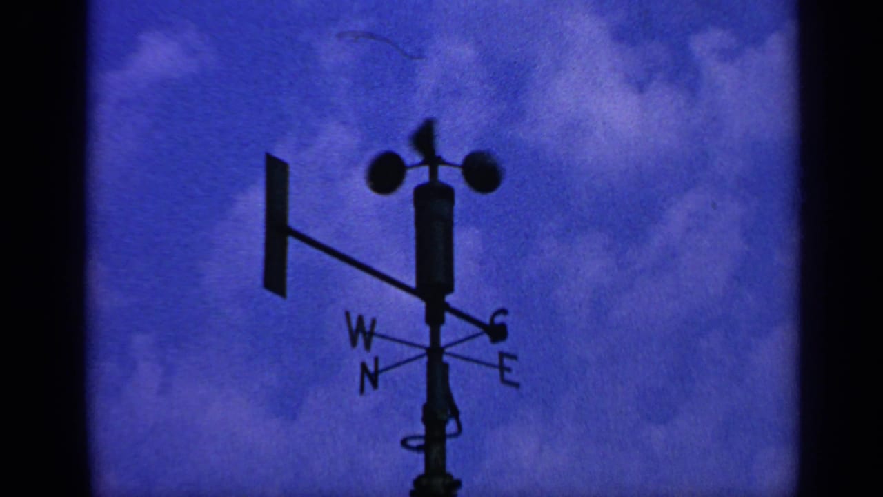1970 United Kingdom 8mm frame showing a silhouetted wind vane and cup anemometer against saturated blue sky and clouds.
