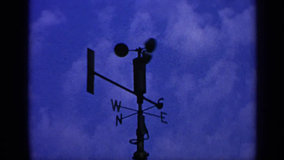 1970 United Kingdom 8mm frame showing the anemometer and vane from below, with grainy blue sky and cloud texture.