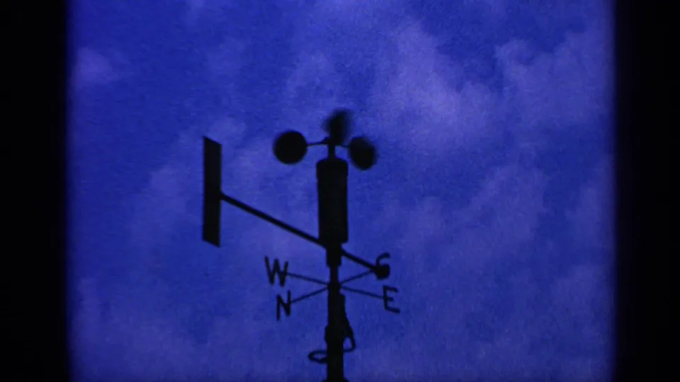 1970 United Kingdom 8mm frame showing a low-angle silhouetted weather vane, cup anemometer, and compass letters against blue cloud cover.