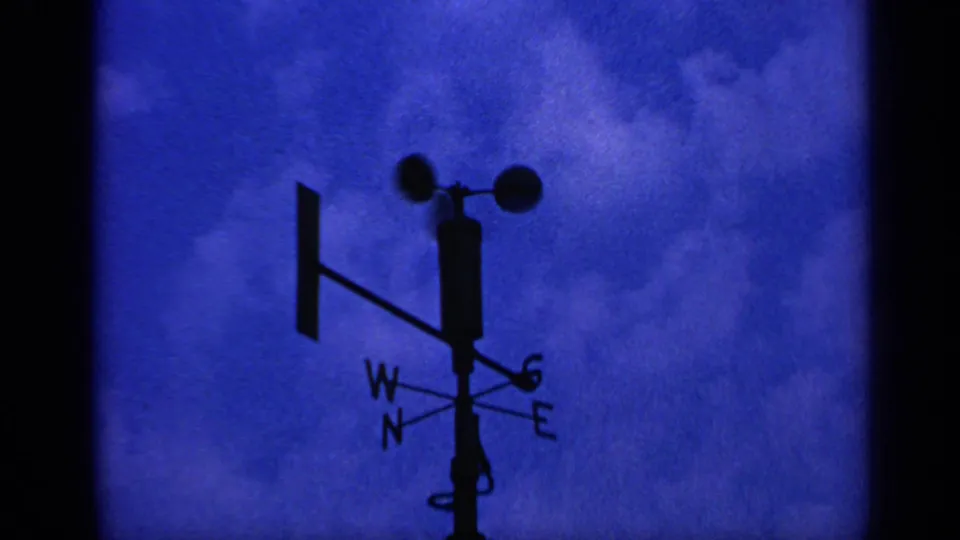 Low-angle 8mm view of a silhouetted weather vane and anemometer in the United Kingdom, set against a saturated blue cloudy sky.