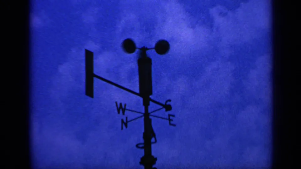 Final 8mm frame showing the cup anemometer, vane, and compass points as a dark silhouette against blue-toned clouds.