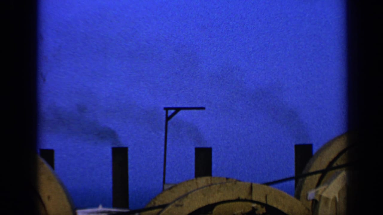 1970 United Kingdom 8mm frame showing dark chimneys, a lamppost, smoke, and curved wooden spools beneath a saturated blue sky.