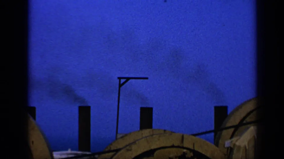 1970 United Kingdom 8mm frame with silhouetted chimneys, a slim lamppost, drifting smoke, and large wooden spools in the foreground.