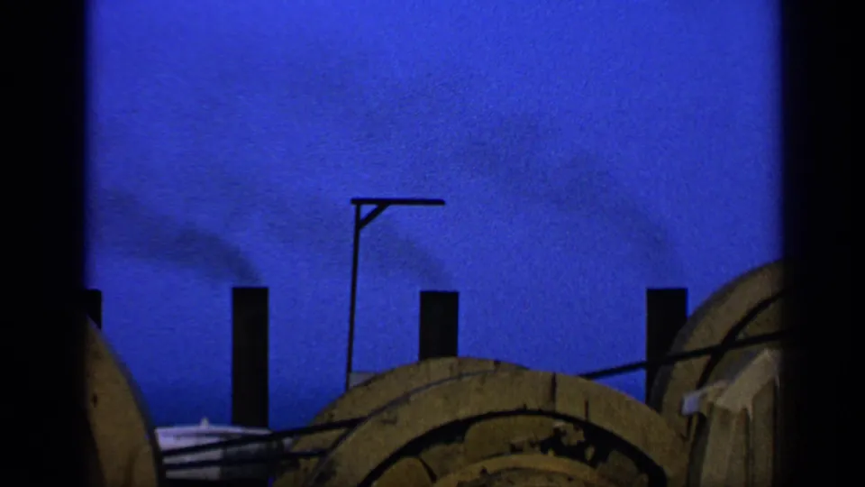 1970 United Kingdom 8mm frame showing smoke near silhouetted chimneys, with a lamppost and broad wooden spool shapes below.