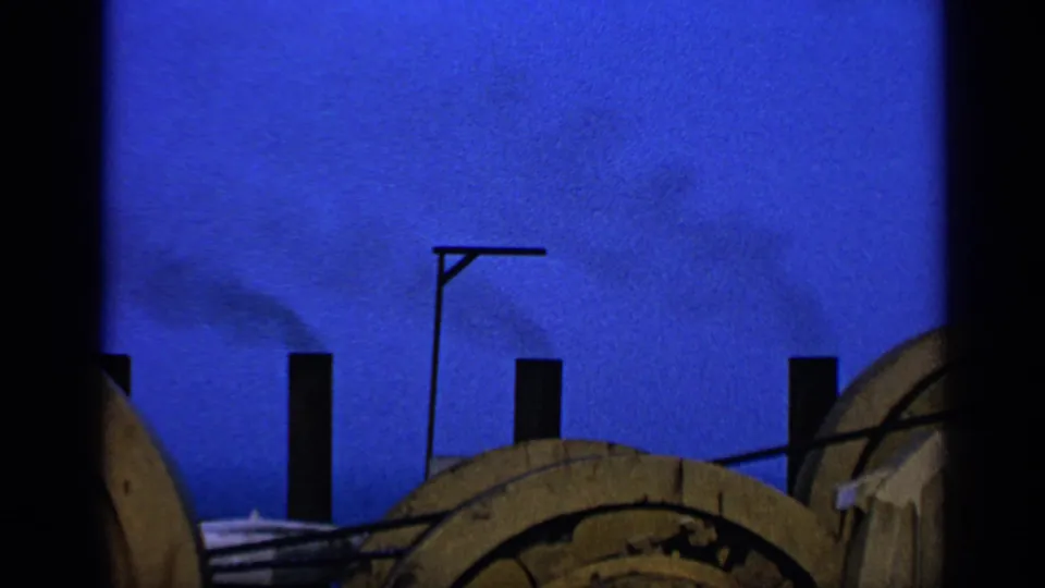 1970 United Kingdom 8mm frame with black chimney silhouettes, drifting smoke, a lamppost, and wooden spools against a vivid blue sky.