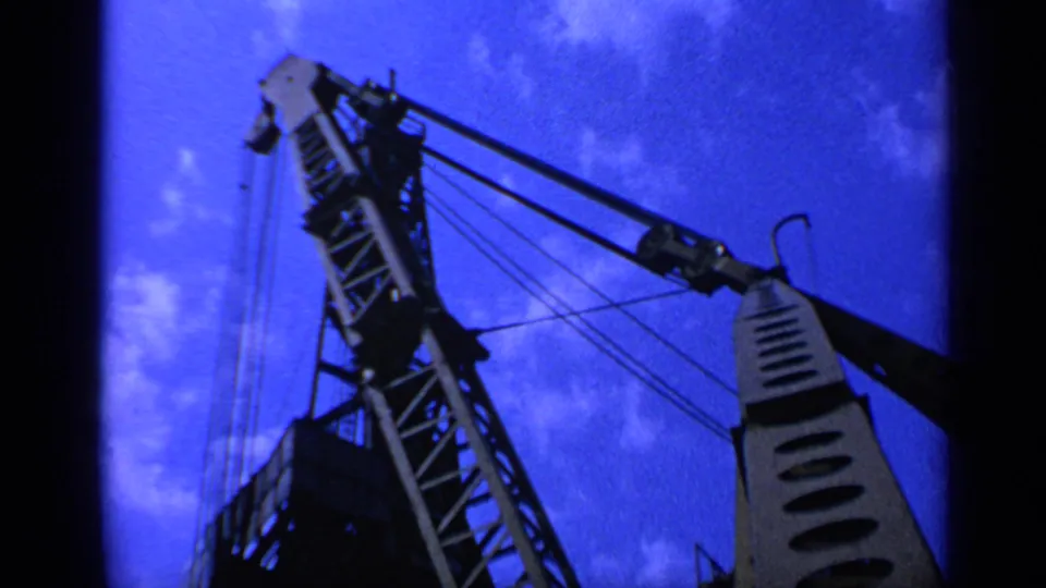 Low-angle 1970 United Kingdom frame of a crane boom, cable rigging, and pulley hardware against blue sky and clouds.