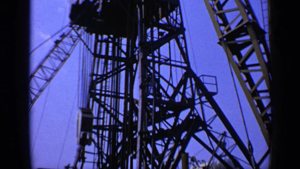 1970s United Kingdom 8mm frame showing a crane tower, diagonal boom, cables, and dark metal framework against saturated blue sky.