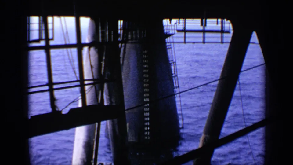 1970 United Kingdom 8mm frame looking through offshore platform railings and legs toward blue ocean water below.