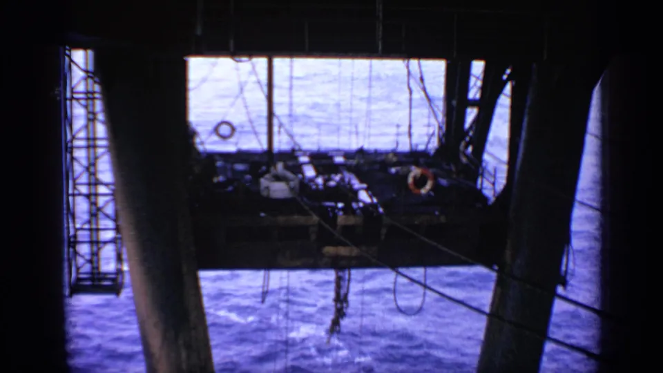1970 United Kingdom 8mm frame looking across an offshore platform deck with railings, cables, ladderwork, a pillar, and blue sea.