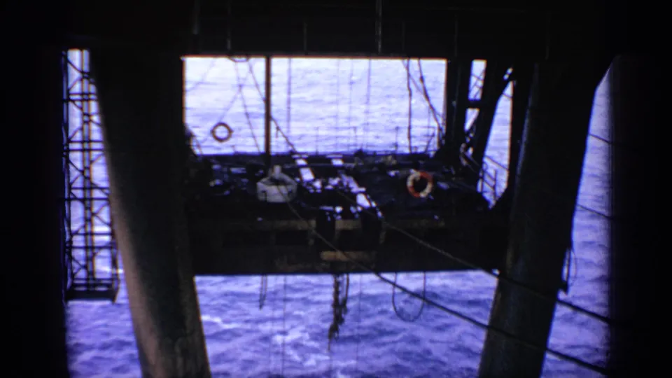 1970 United Kingdom frame showing a shadowed offshore deck, heavy pillar, ladder sections, cables, and the surrounding blue ocean.