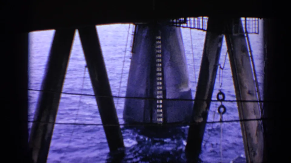 Obstructed 1970 United Kingdom offshore rig view with crossed supports, water below and hanging industrial fittings.