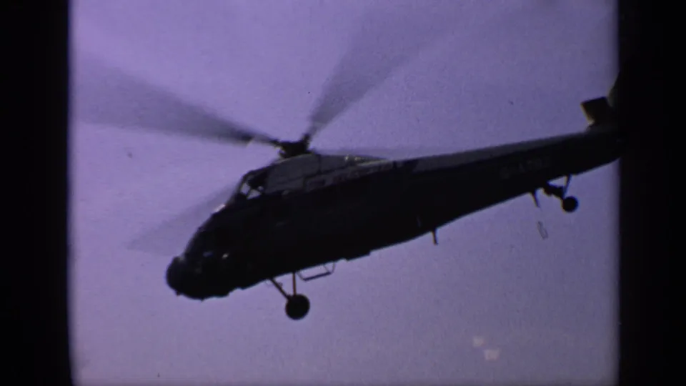1970 United Kingdom 8mm frame of a dark helicopter flying low against a hazy sky, with rotor blur and partial registration or markings visible.