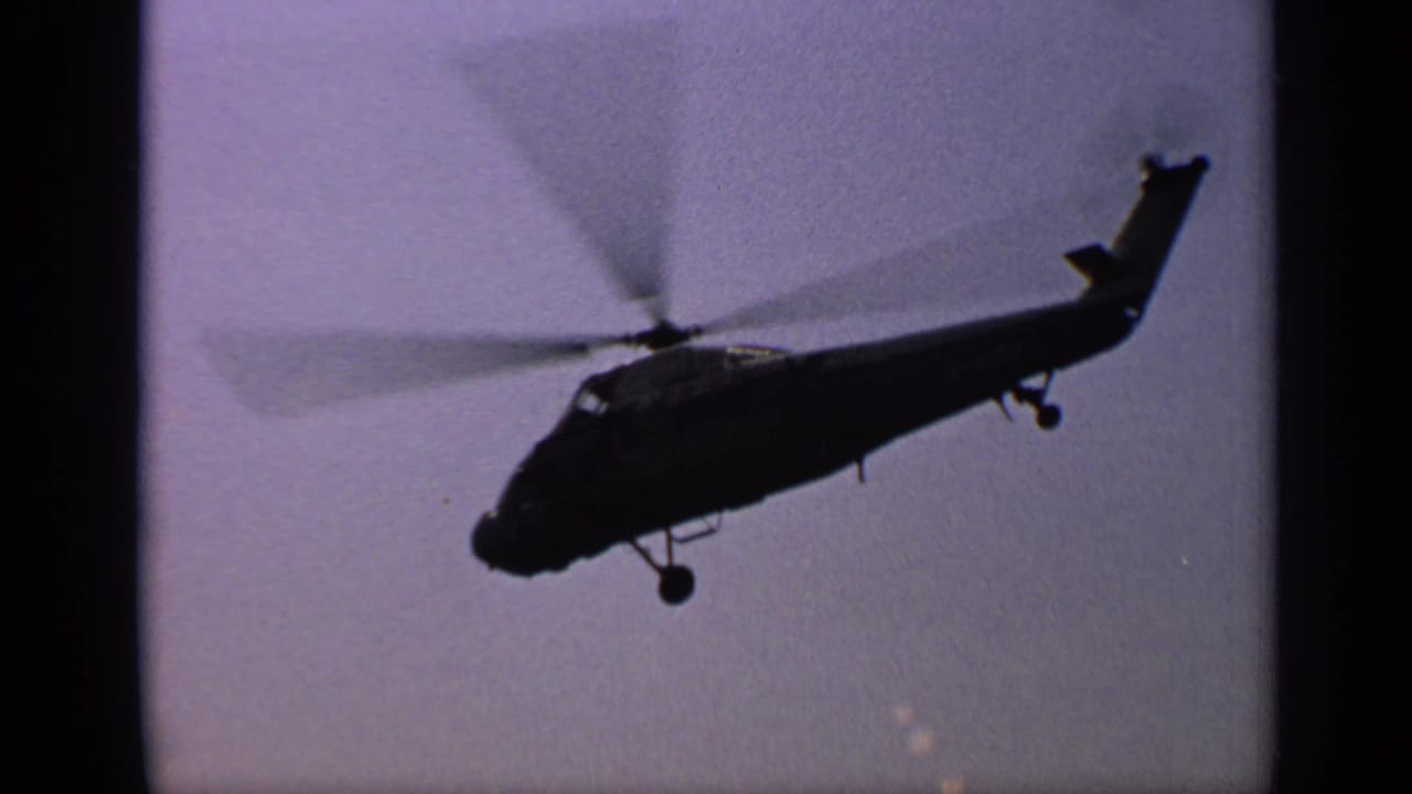 Helicopter silhouette flying across a pale sky with spinning rotor and landing wheel visible.