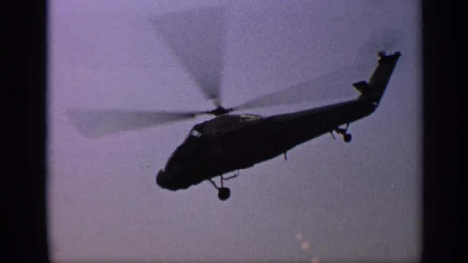 1970 United Kingdom 8mm frame showing a helicopter in silhouette against a pale sky, with rotor blur, tail, and landing gear visible.