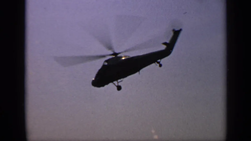 1970 United Kingdom 8mm frame of a helicopter viewed from below in a hazy sky, showing the tail boom, spinning rotor, and landing gear.