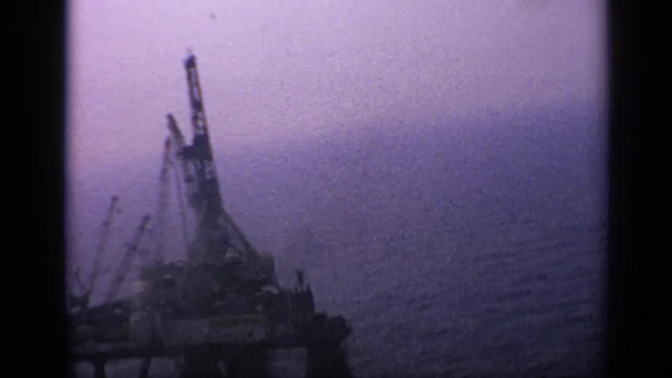 1970 United Kingdom 8mm frame showing a distant offshore rig or industrial vessel with cranes above the sea in a hazy purple frame.