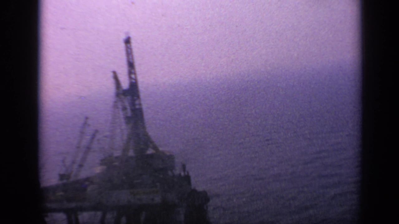 1970 United Kingdom 8mm frame showing a distant offshore rig or industrial vessel in hazy low light over open ocean.