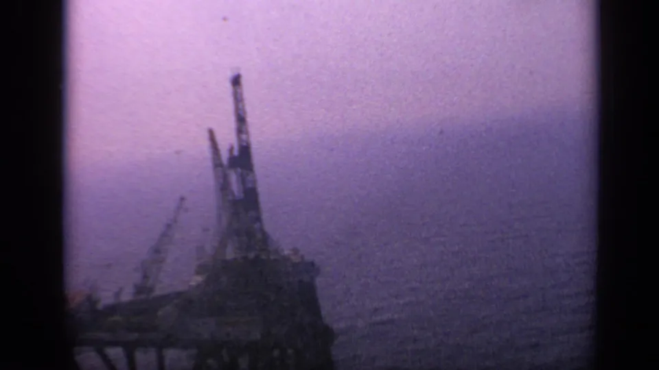Distant offshore rig or industrial vessel appears in silhouette on 1970 United Kingdom 8mm footage, with calm open water behind it.