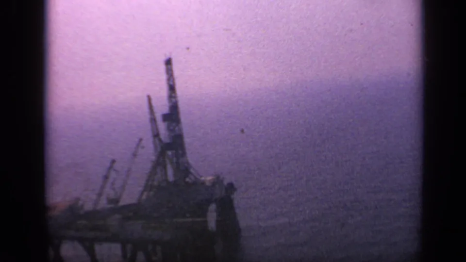 1970 United Kingdom 8mm frame with a dark offshore industrial structure, visible crane shapes, and a hazy sea horizon.