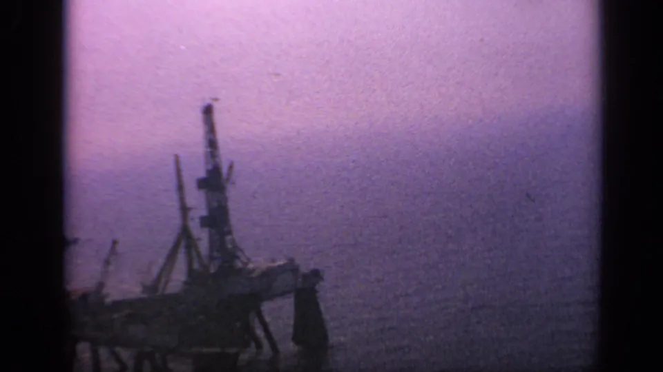 1970 United Kingdom 8mm frame showing a silhouetted offshore platform on the ocean under purple low-light haze.