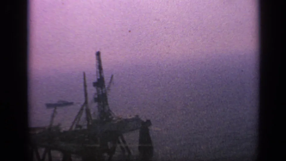 1970 United Kingdom 8mm frame with a dark offshore platform and a faint ship-like form in the background haze.