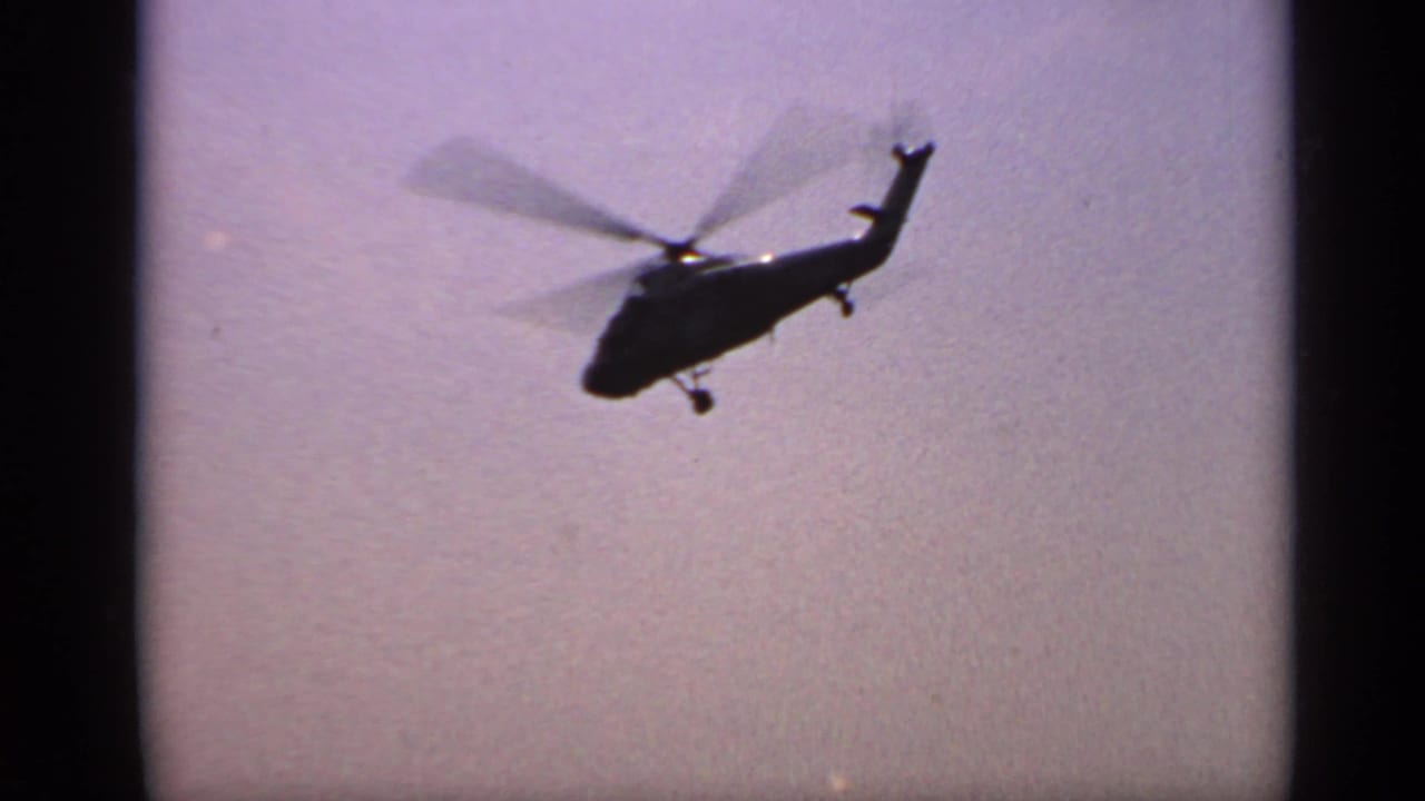 A dark helicopter silhouette flies across a hazy sky above open water.