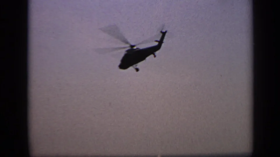 1970 United Kingdom 8mm frame showing a helicopter in flight, dark against the misty sky above water.