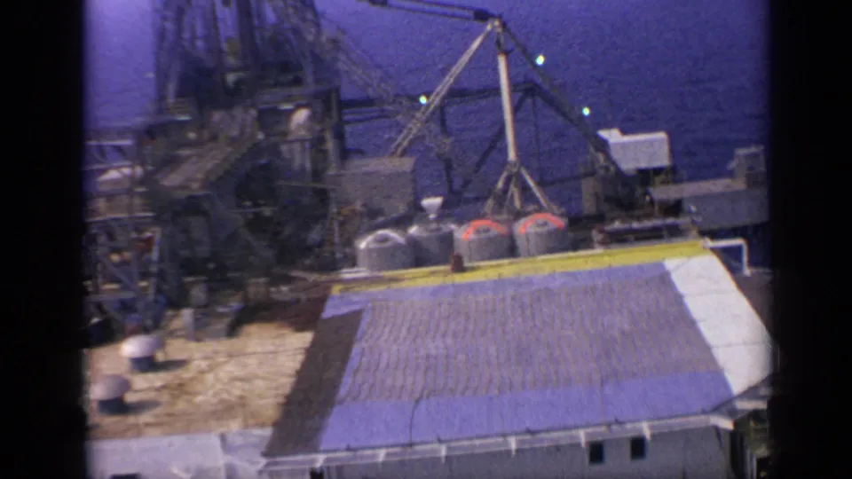 A soft-focus 8mm view shows the rig deck, roofline, tanks, crane structure, and ocean backdrop in 1970 United Kingdom footage.