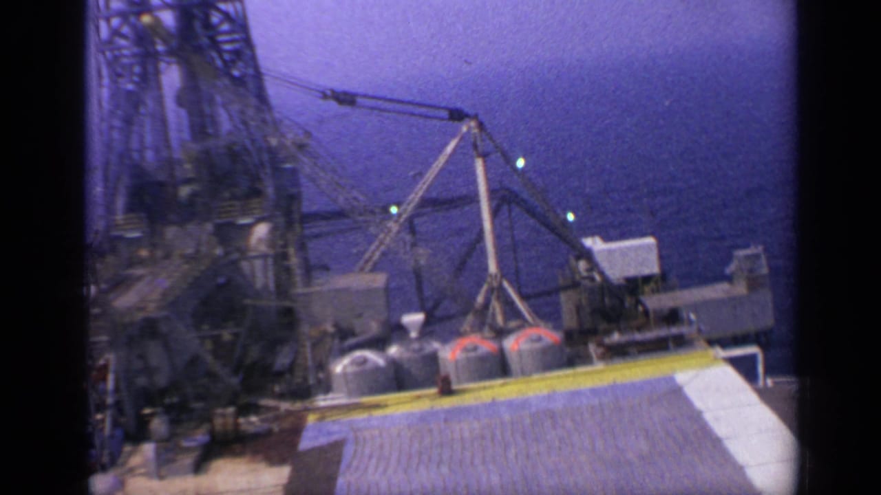 Offshore platform deck with derrick, crane boom, tanks, and ocean beyond.
