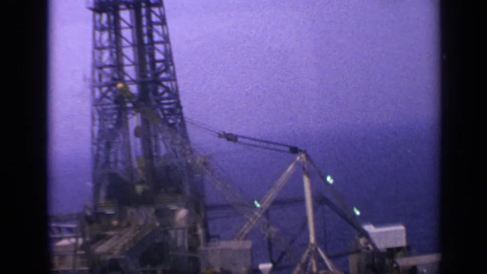 The camera frames the derrick and crane rigging on an offshore oil platform, with a purple dusk sky and ocean horizon behind it.