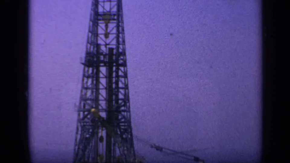 Upward 8mm frame of the offshore derrick structure, showing dark latticework and crane rigging against a grainy purple sky.