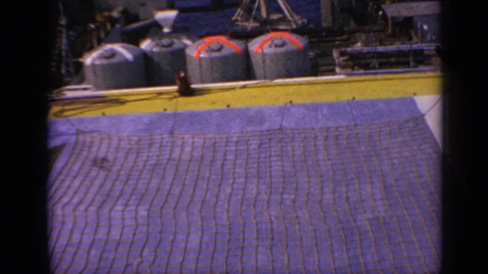 1970 UK 8mm frame showing a platform deck, brown net or mat, yellow edge and industrial tanks in the background.