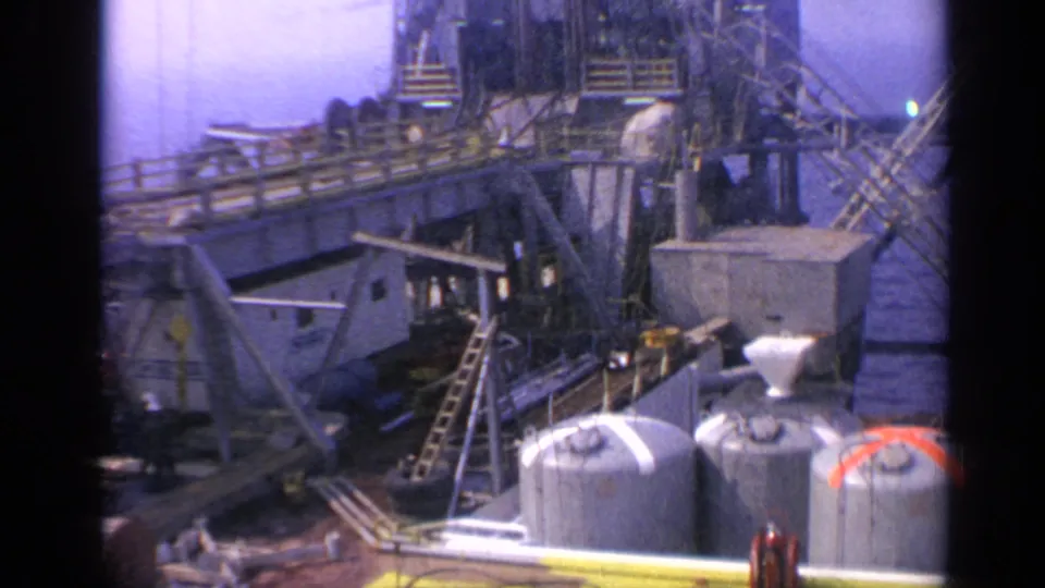 1970 United Kingdom 8mm frame of an offshore platform deck with foreground tanks, yellow edging, stairs, ladders, and dense rig structure.