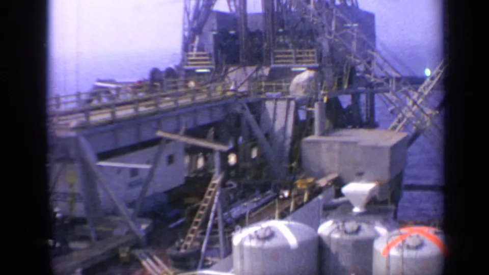 1970 United Kingdom 8mm frame showing oil-rig equipment, storage tanks, walkways, ladders, and industrial structures under a bright sky.
