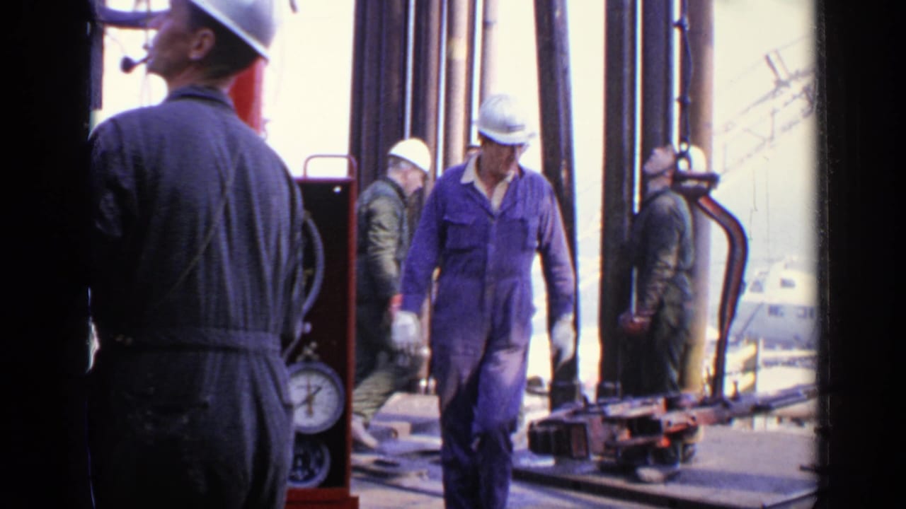 Hard-hatted workers in coveralls stand and walk among pipes and machinery on a rig platform.
