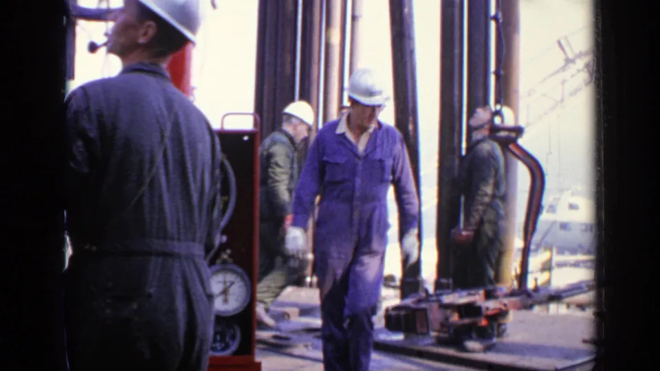 1970 United Kingdom frame of hard-hatted oil-rig workers in coveralls moving near large vertical pipes and platform machinery.