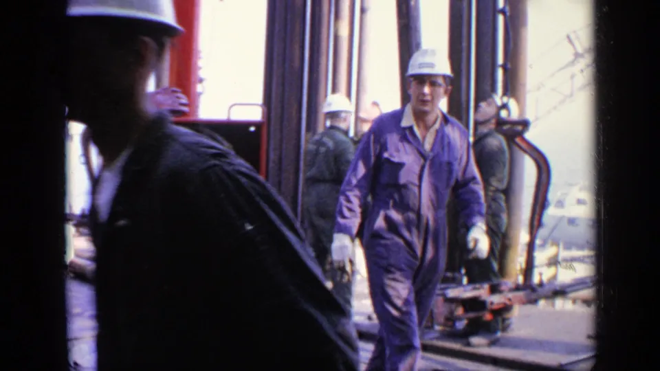 1970 United Kingdom 8mm frame with a worker in purple coveralls approaching camera as others stand near rig pipes and equipment.