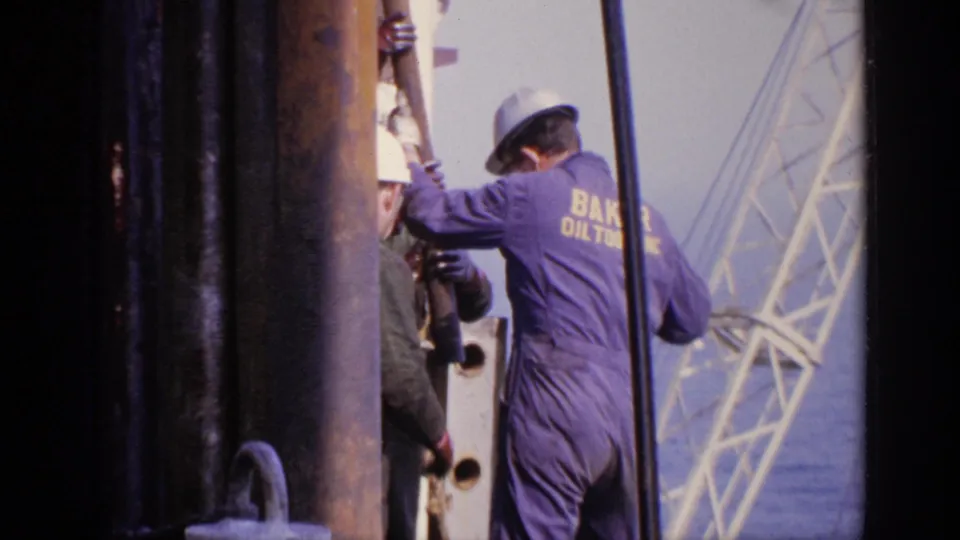 1970 United Kingdom frame showing a hard-hatted worker with Baker Oil Tools on his coveralls beside large pipes and rig structures.