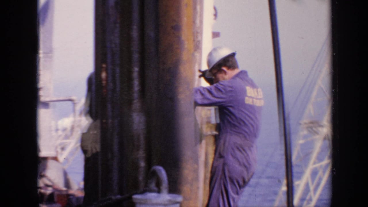 Worker in a purple BAKER OIL TOOLS coverall beside vertical pipes on an offshore platform.