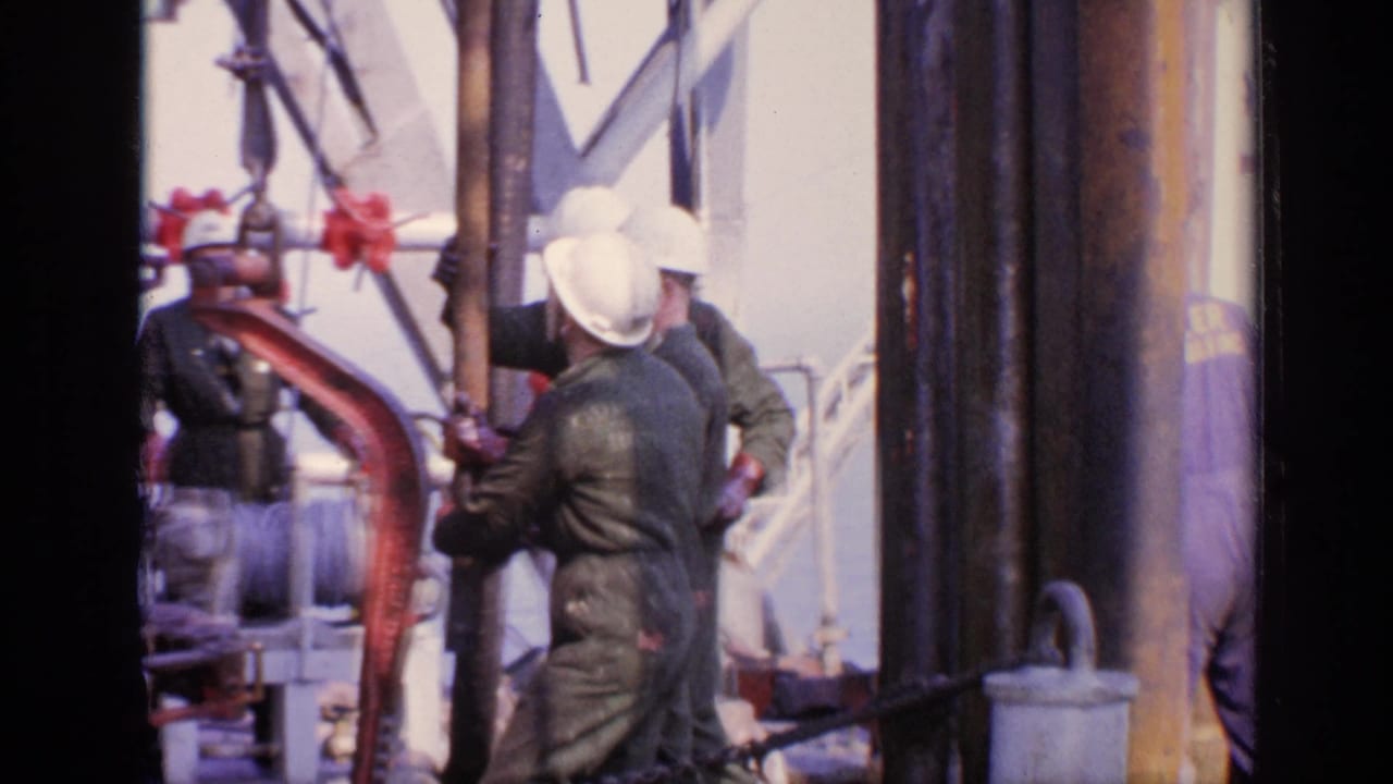 Hard-hatted workers gather around red deck machinery and vertical pipework on an offshore platform.