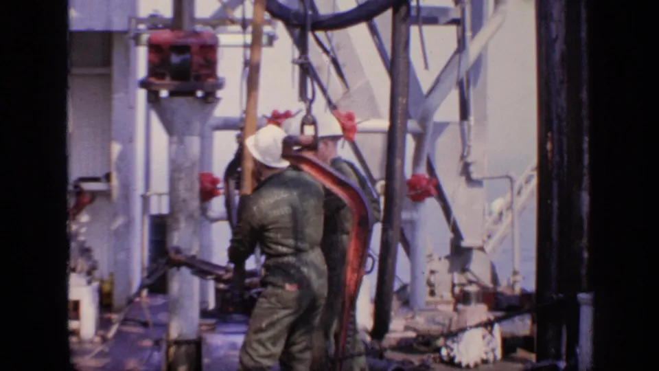 1970 United Kingdom 8mm frame showing a worker in dark coveralls near red machinery, pipes, and metal structures on an offshore platform.